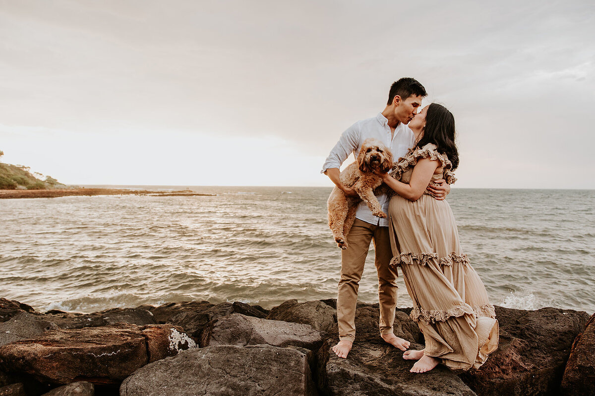 Man and woman kiss while standing on rocks at the beach with their dog in foreground at Melbourne maternity session
