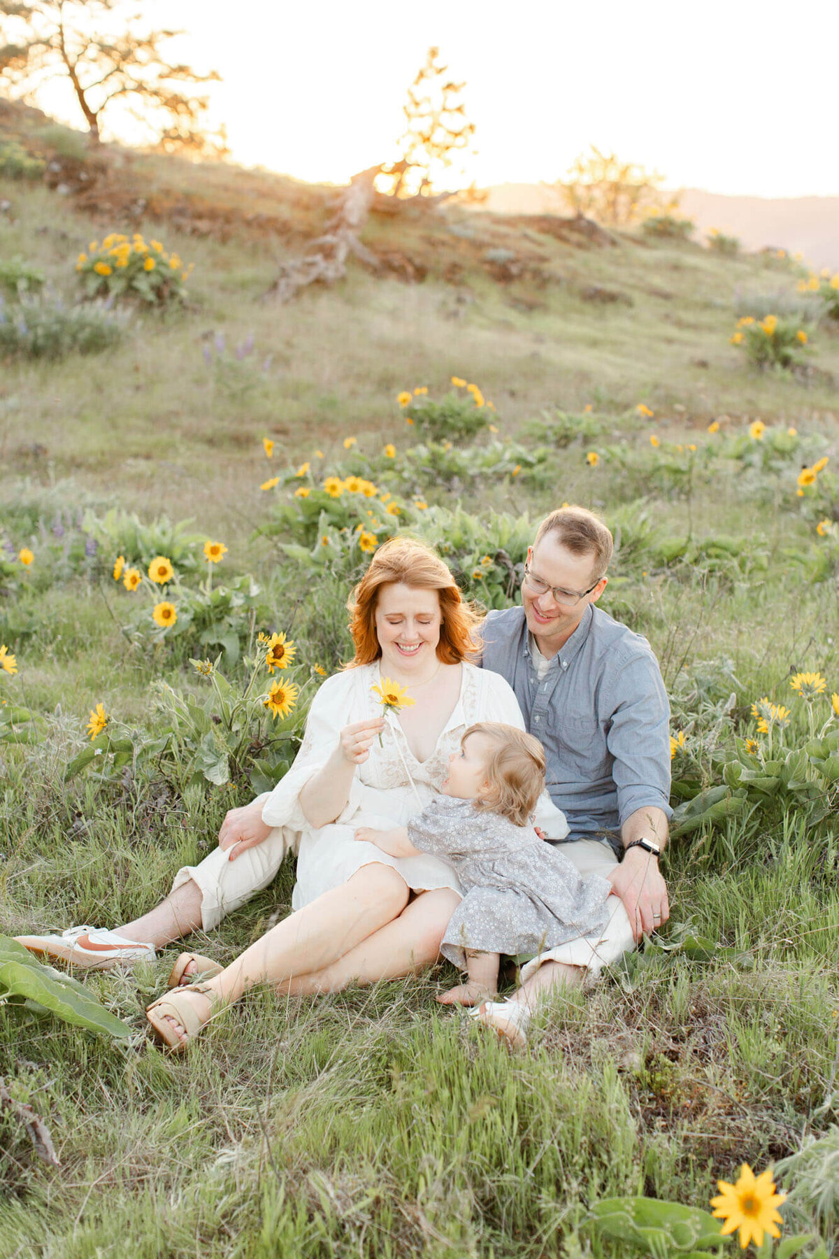 Mom, Dad and one year old girl sitting in a field of yellow wildflowers together. Mom is holding up a flower and little girl is looking up at it and smiling. Dad is sitting behind them and smiling at them. They are wearing shades of white, blue and grey.
