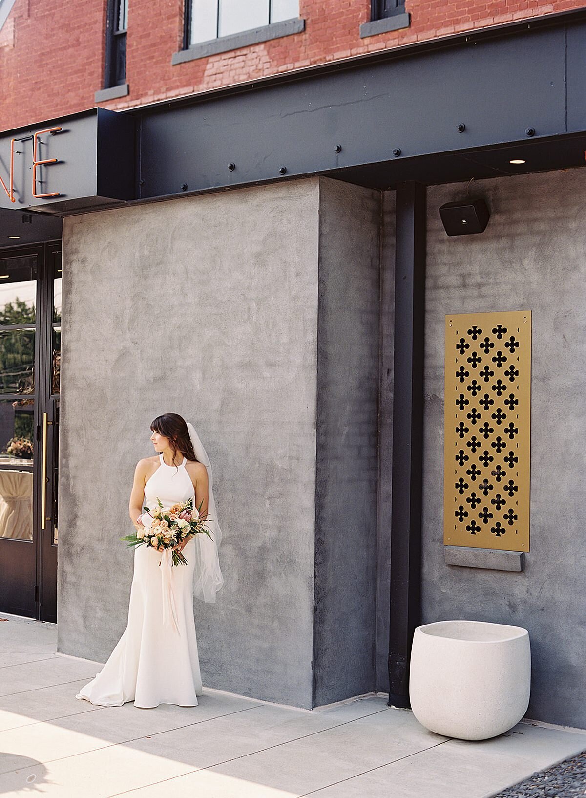 The bride, wearing a halter top silk sheath wedding dress and long veil stands in the courtyard at Clementine Hall holding her horizontal cascade bouquet filled with white, ivory, peach, yellow and pink tropical flowers, orchids, palm fronds and protea. The courtyard of Clementine Hall is accented with gold filigree windows, soft grey walls, white planters and glass doors.