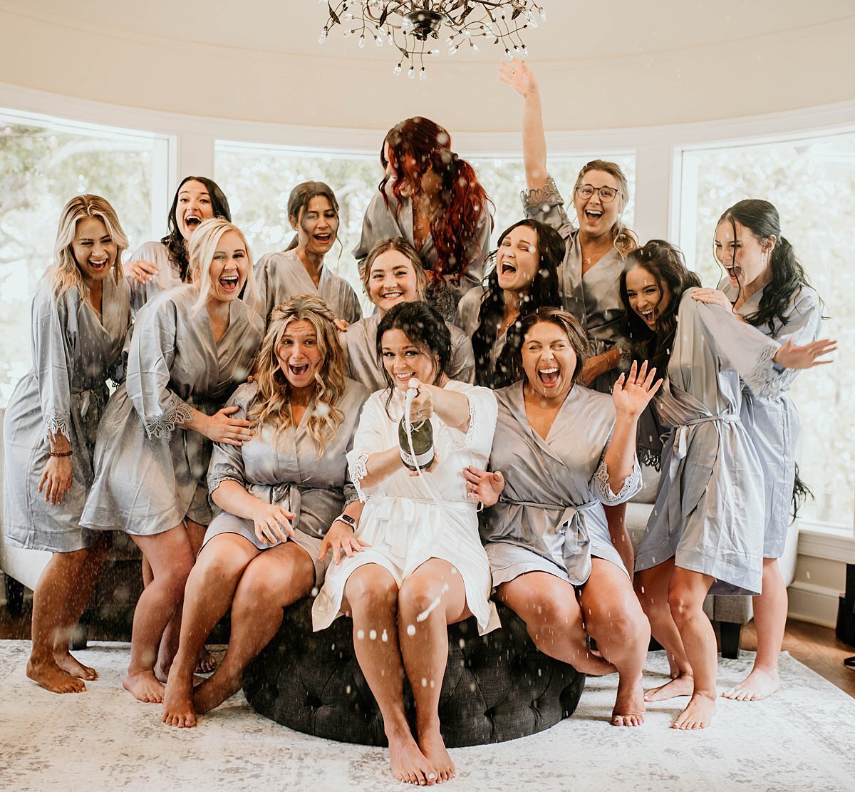 A bride wearing a white silk robe sitting on a black ottoman pops a bottle of champagne as her twelve bridesmaids in matching silk silver robes. The bridesmaids cheer on the bride at The Estate at Cherokee Dock for her Nashville Wedding.
