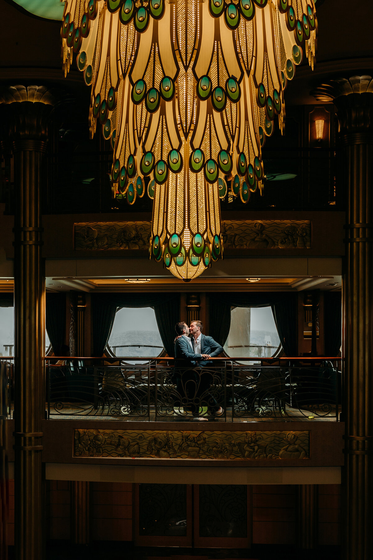 Two grooms share a kiss beneath the peacock chandelier aboard the Disney Fantasy during their cruise ship wedding portraits.