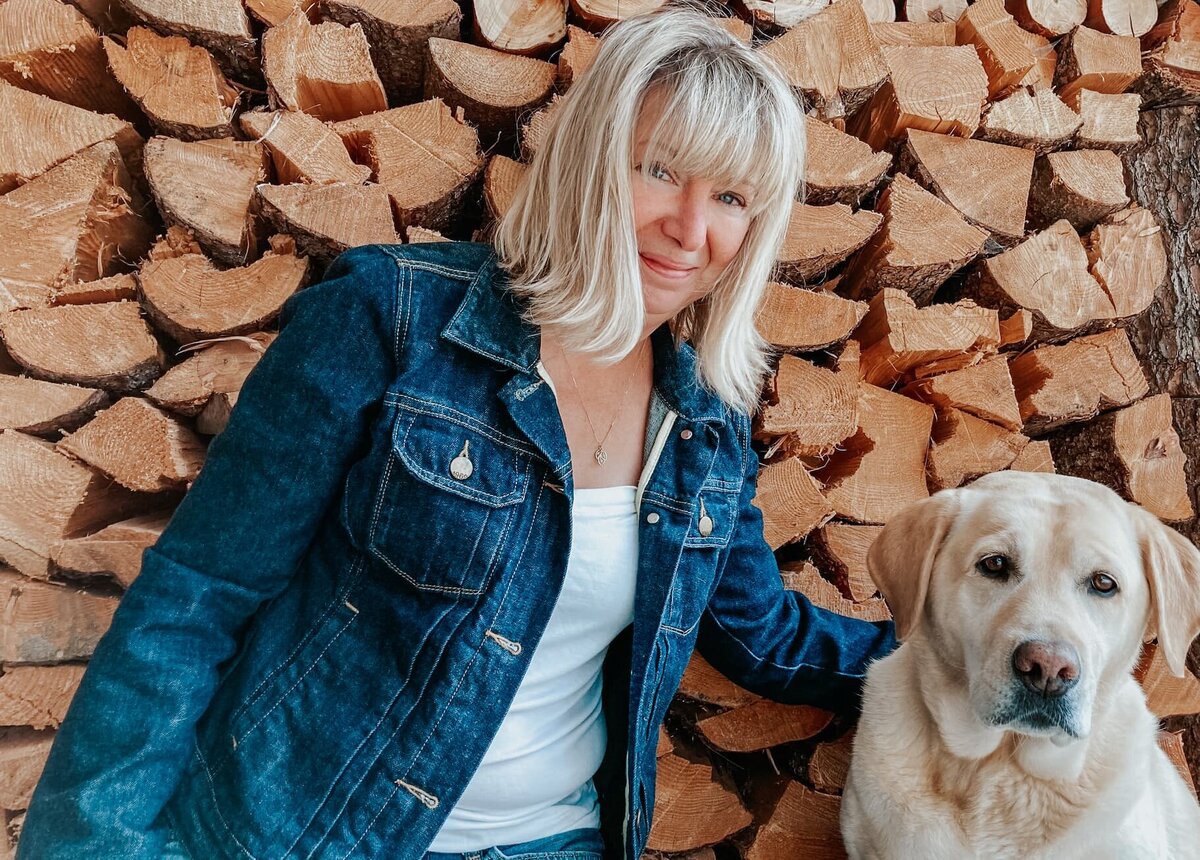 Susan Wyatt sitting with her Labrador, Flora, in front of stacked firewood, representing her expertise in landscape design and plant propagation.