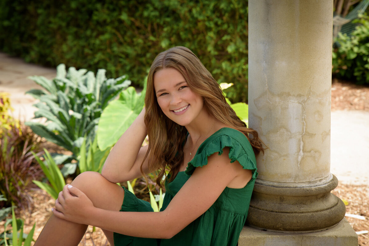 Luxemburg Casco High School senior girl wearing short kelly green dress sitting by a cement pillar at the Green Bay Botanical Gardens in Green Bay, Wisconsin