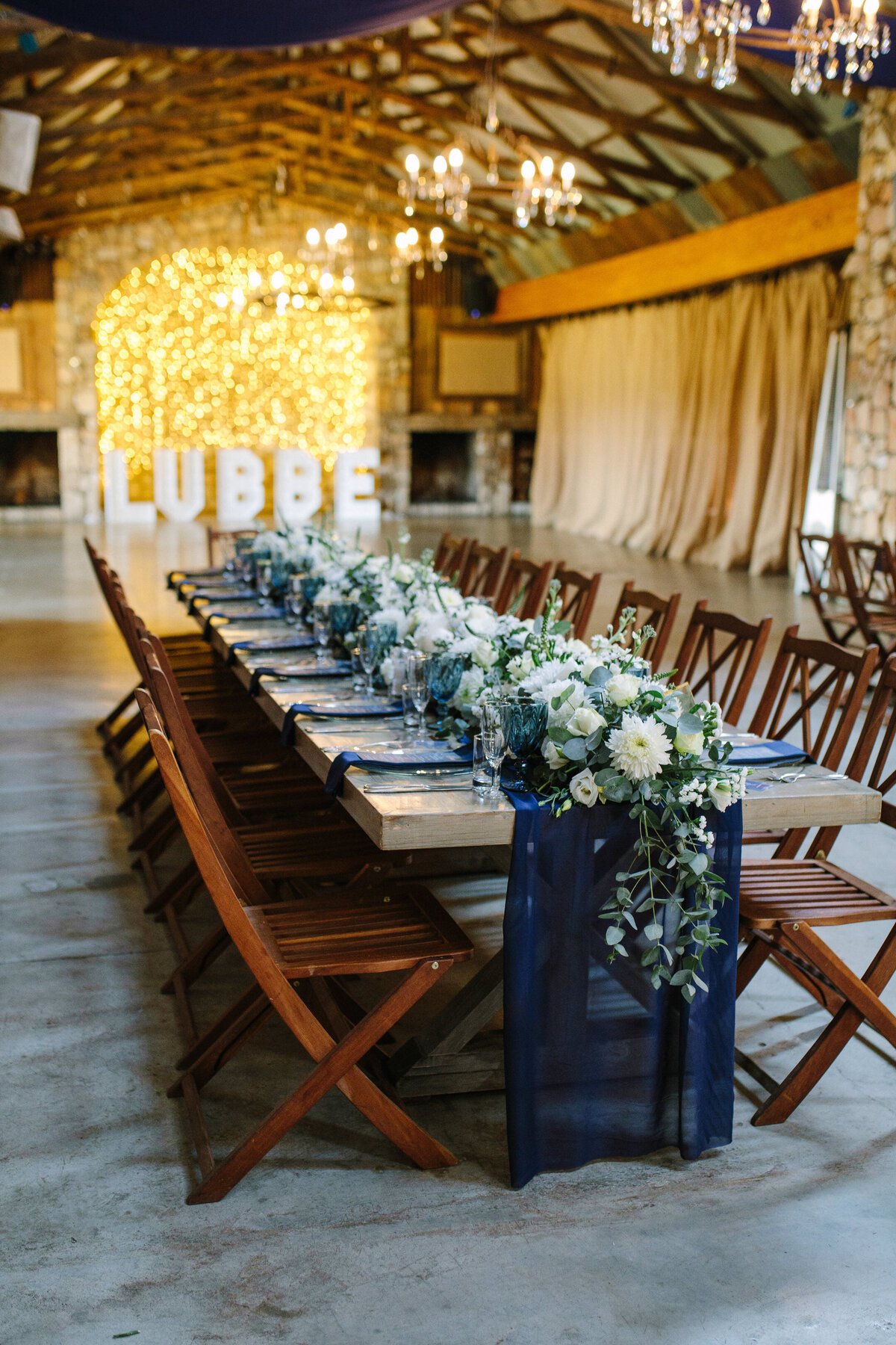 Elegant table setting at a wedding reception featuring navy décor, flowing floral arrangements, a candle centerpiece, and twinkling fairy lights behind.