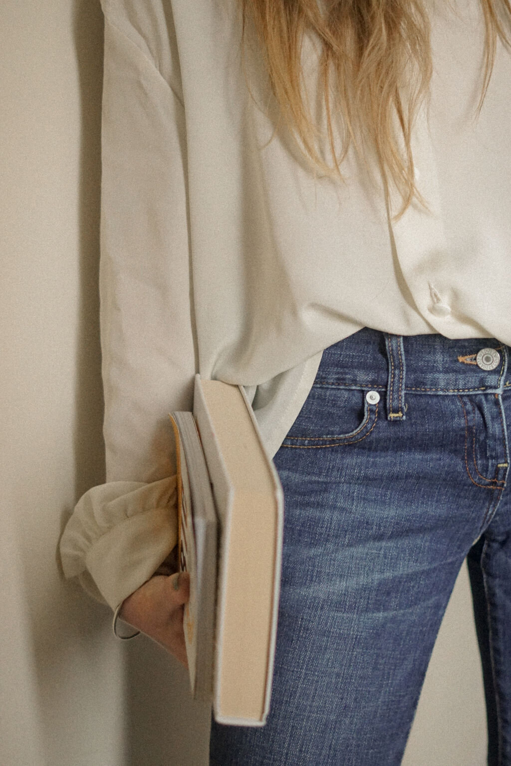 A close-up of a woman with long blonde hair, dressed in an oversized cream silk blouse and jeans, holding a stack of books, embodying a refined yet relaxed style.