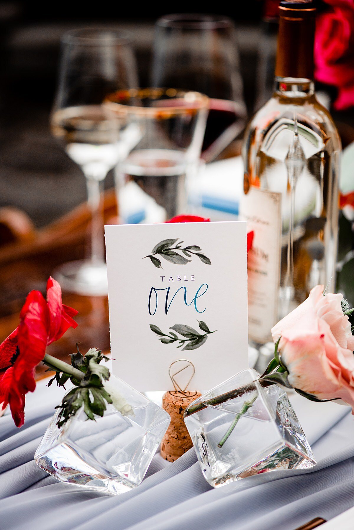 The hand drawn table number card with gray watercolor leaf vines and a teal hand calligraphy ONE is sitting in a wine cork stand between two ice cube glass bud vases with a red rose and a blush rose sitting on top of a dove gray organza tale runner with a bottle of Arrington Vineyards white wine and two wine glasses in the background.