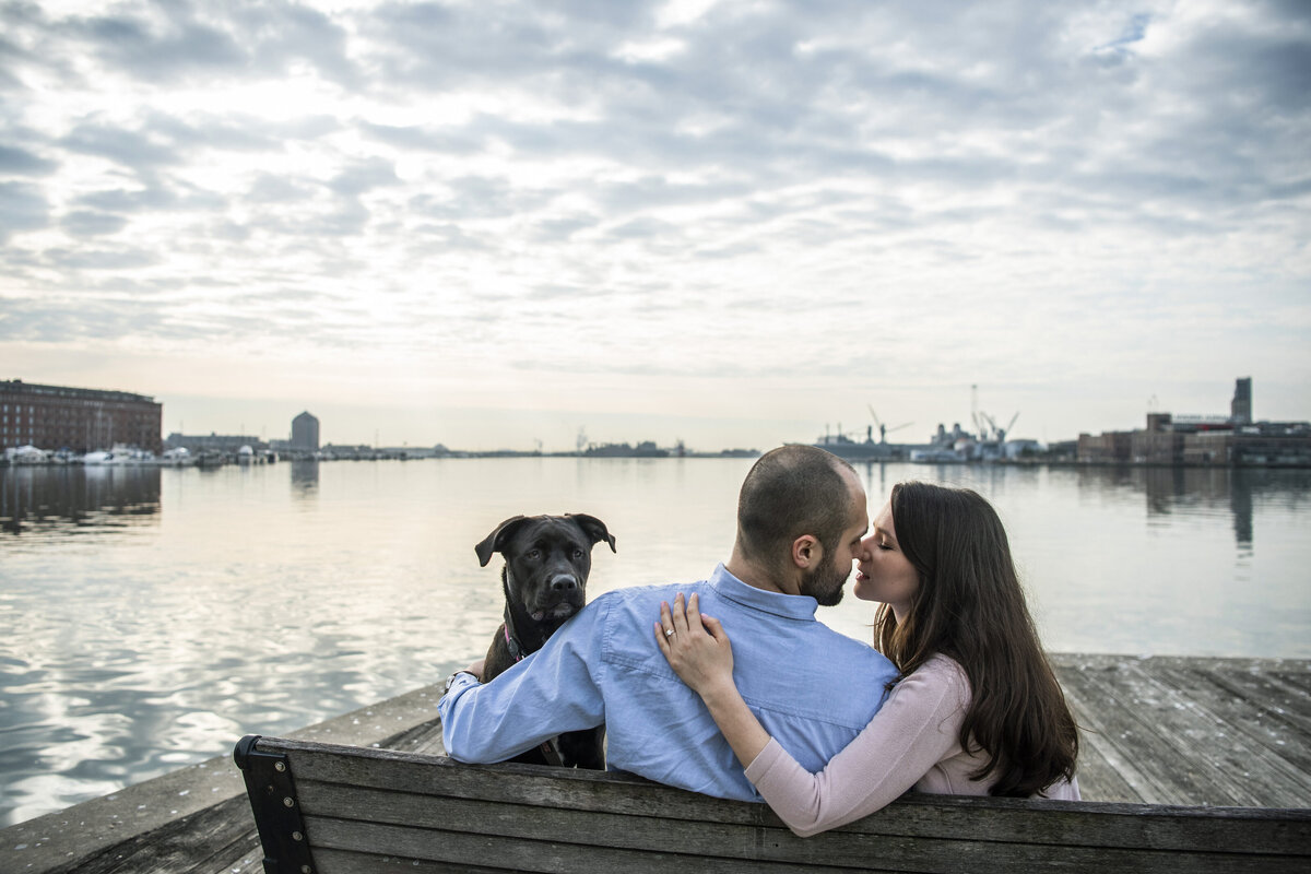 Two people lean in for a kiss in front of the water, their dog with them.