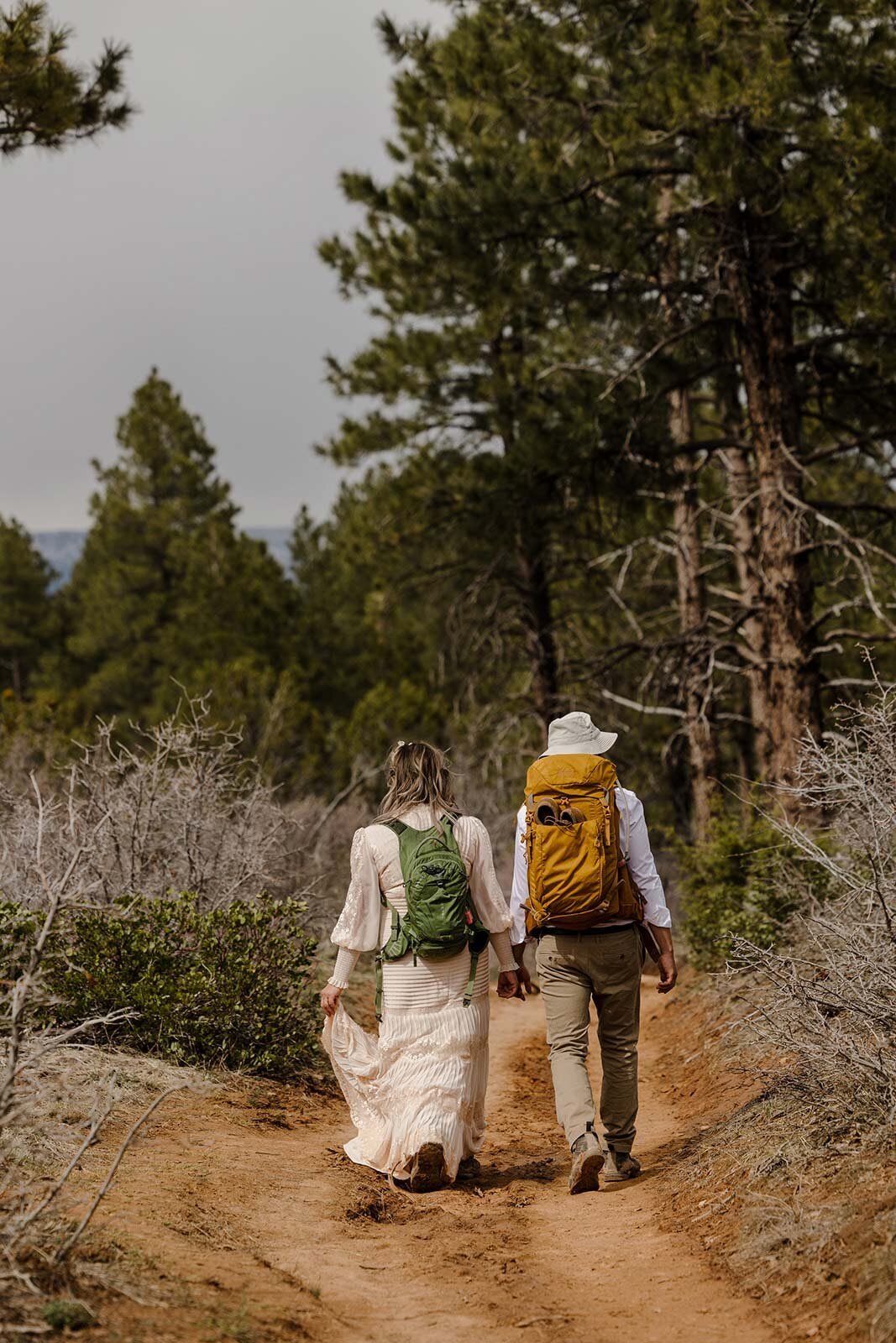 A bride and groom hiking on the trail. They are both in their wedding attire and backpacks. 