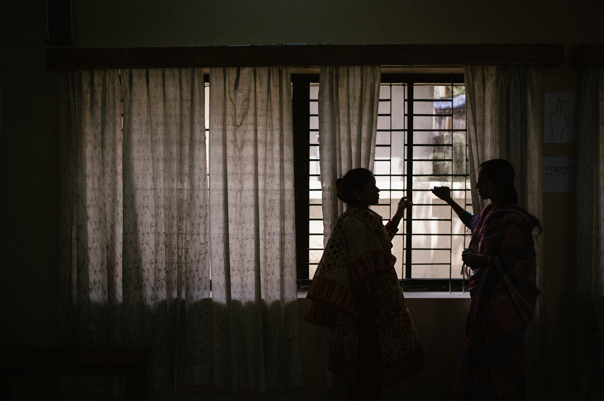 vrouwen staan voor het raam waardoor je alleen hun silhouet kan zien. Het licht valt door de lamellen heen naar binnen