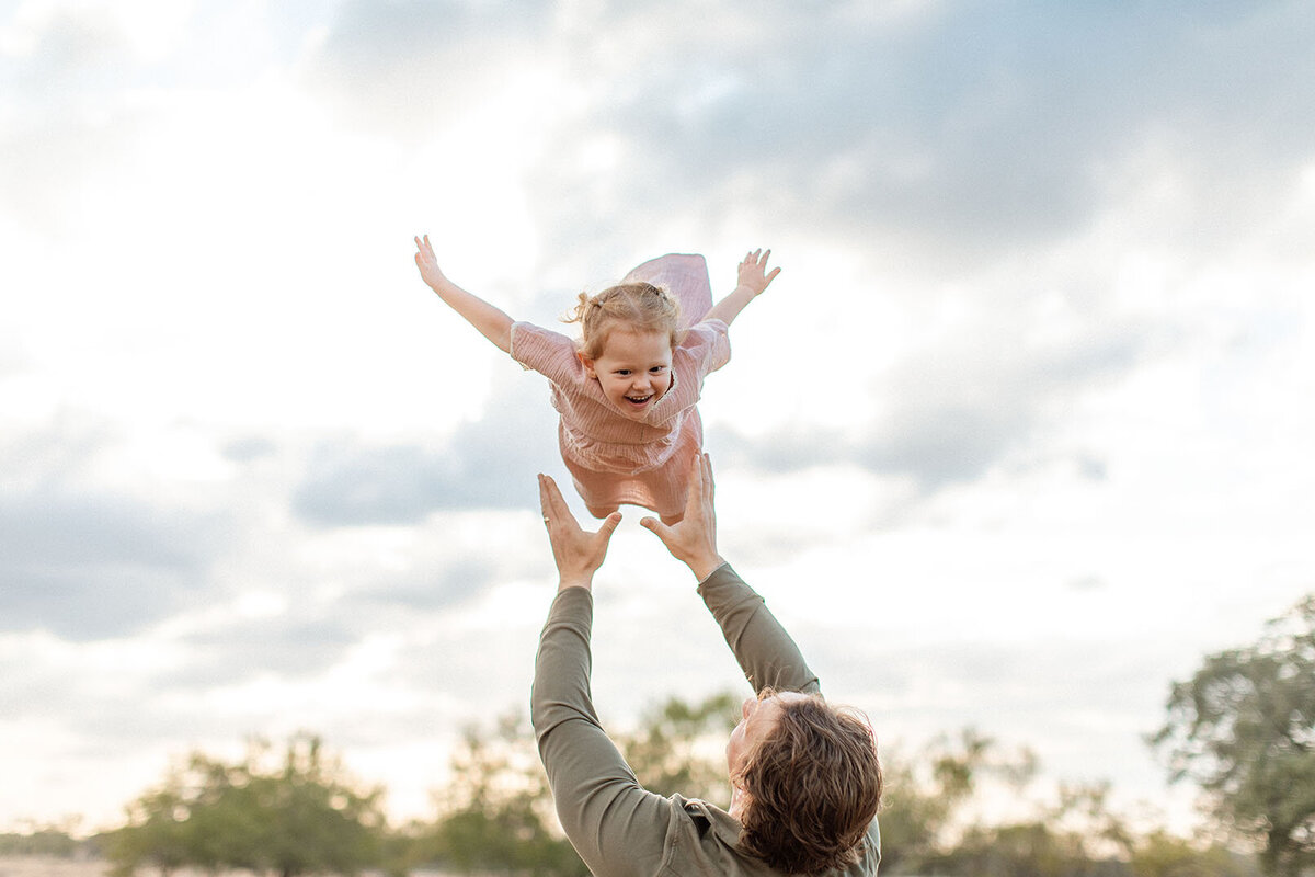 Father throwing daughter in air with beautiful golden light - Sara James San Antonio photographer captures playful father-daughter moments and joyful family interactions in Texas outdoor settings
