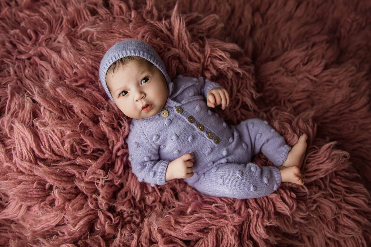 : A newborn baby lies on a plush, textured pink blanket, wearing a lavender outfit with a matching knitted hat. The baby has a curious expression, with hands raised slightly and legs relaxed, capturing a serene moment.