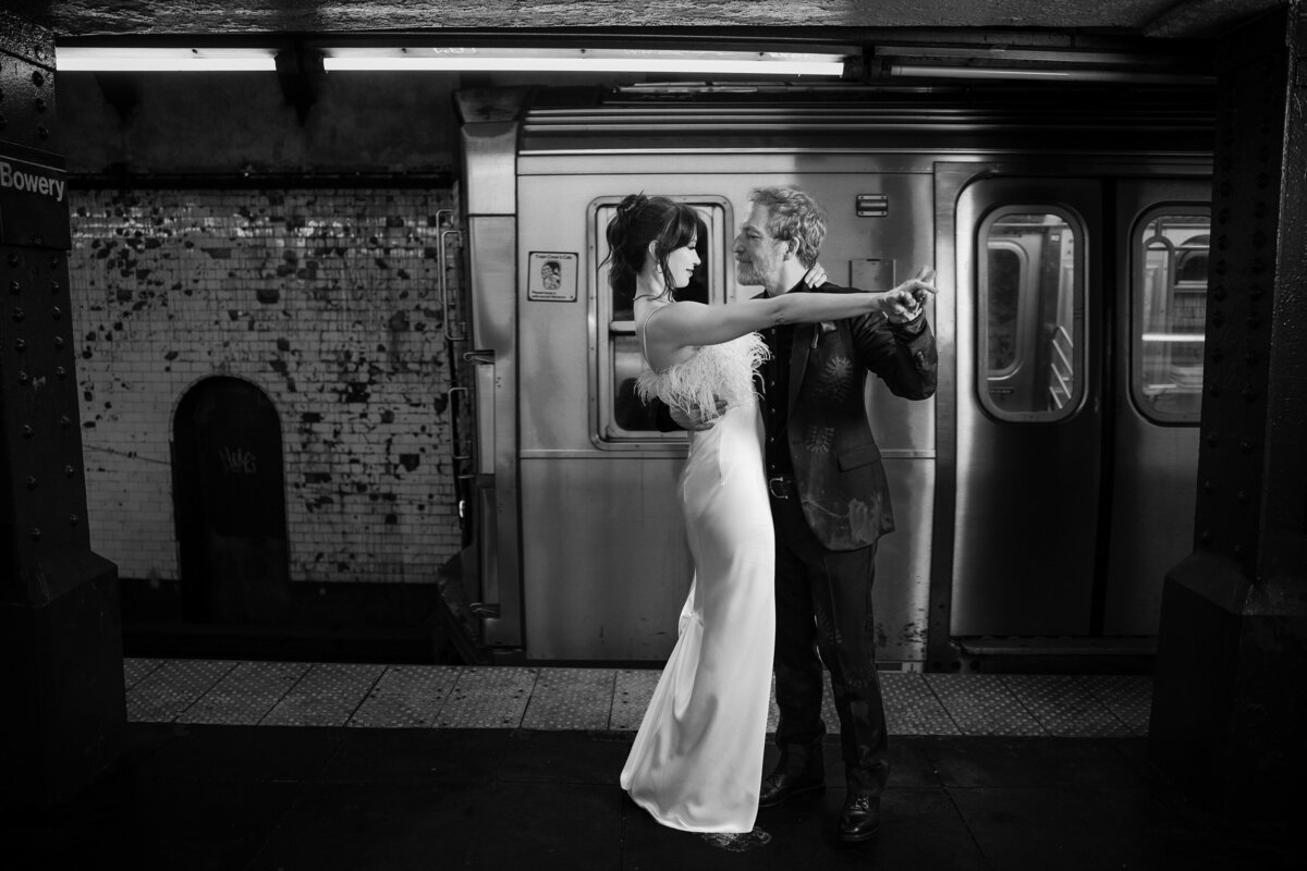 A black-and-white photo of a couple in wedding attire in front of a moving subway train in New York