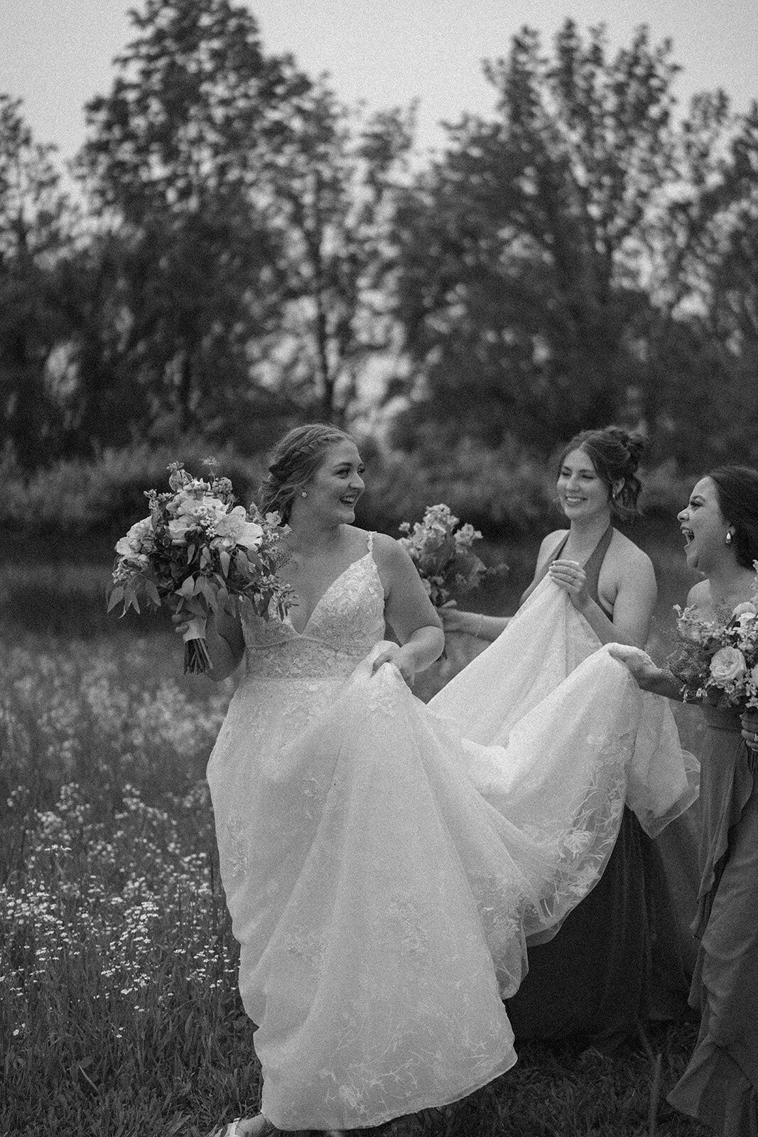 Fun candid moment of bridesmaids holding the bride’s dress while walking and laughing at The Blue Heron Barn wedding.