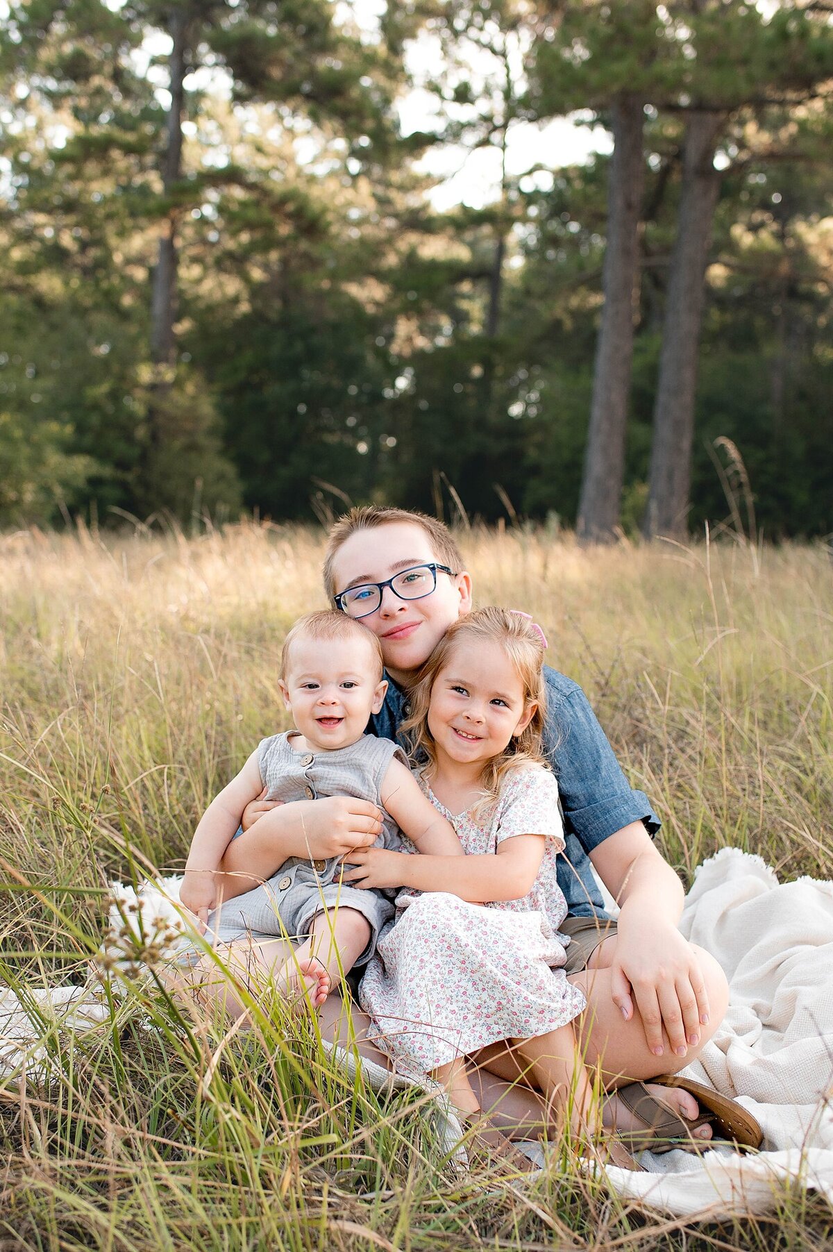 3 kids sitting on blanket in field smiling by Cypress Family Photographer