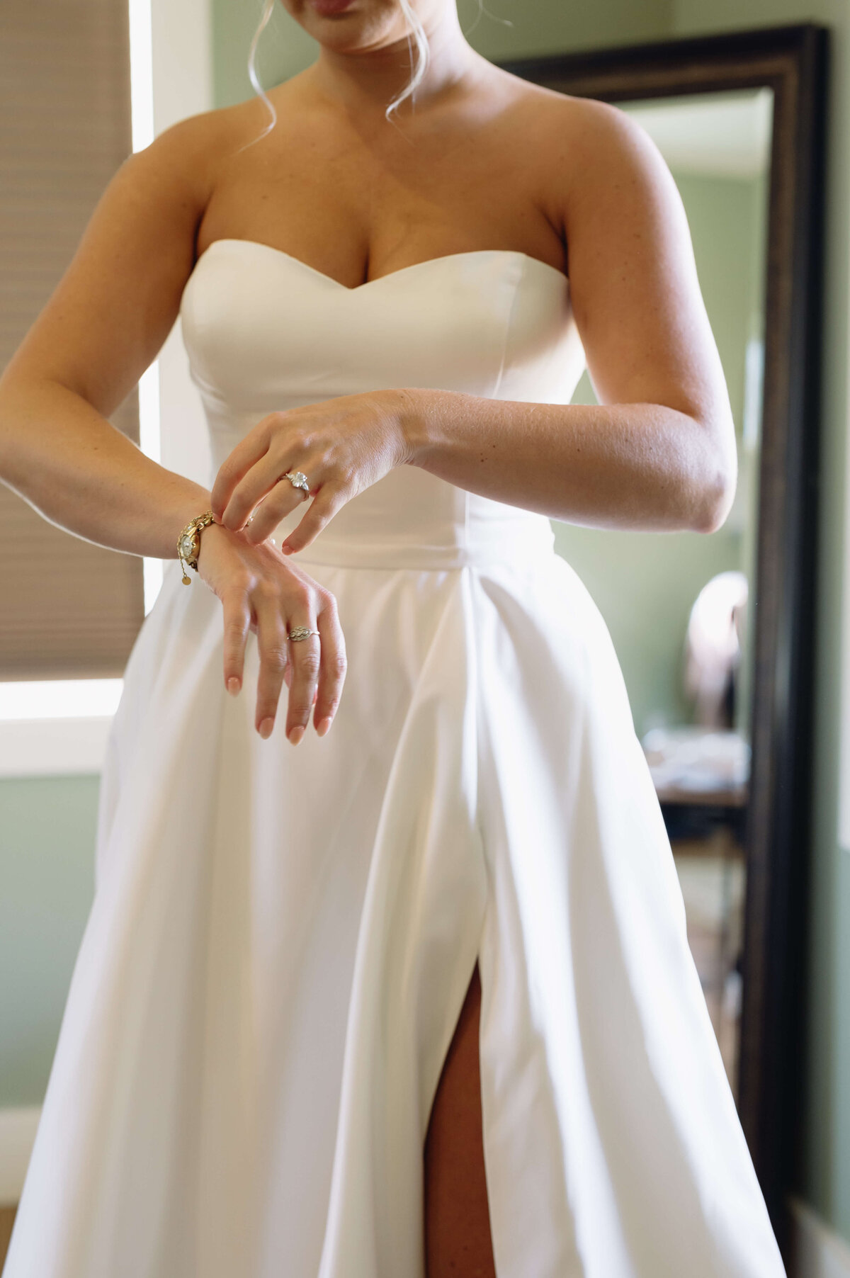 detail photo of bride putting on her jewelry in her bridal suit after she has her gown on qith the light from the window shining in on her