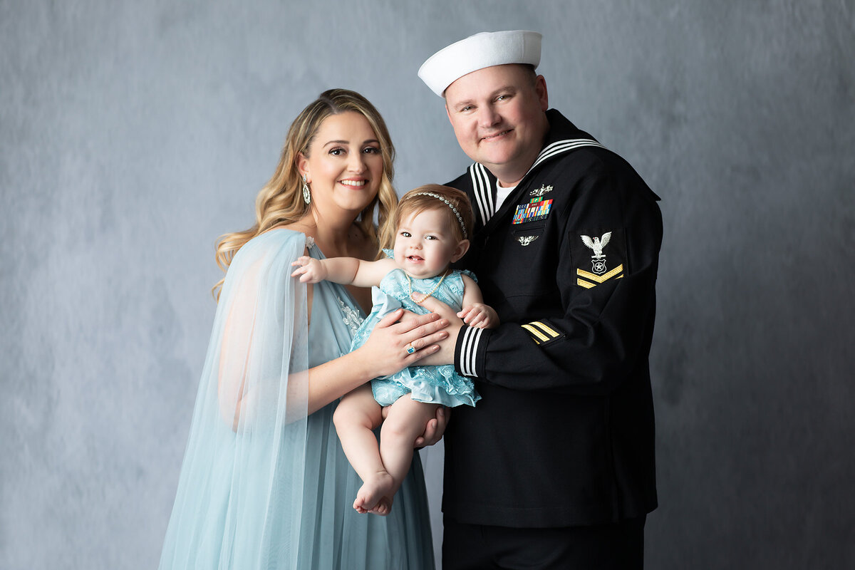 Smiling family poses with their baby girl in a light blue dress as her father wears a Navy uniform, captured during a DFW milestone photography session.