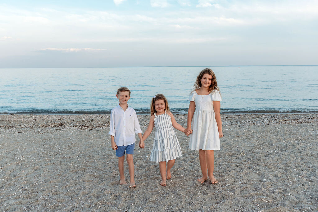 Three children holding hands on beach during sunset captured by Connecticut family photographer in natural coastal setting