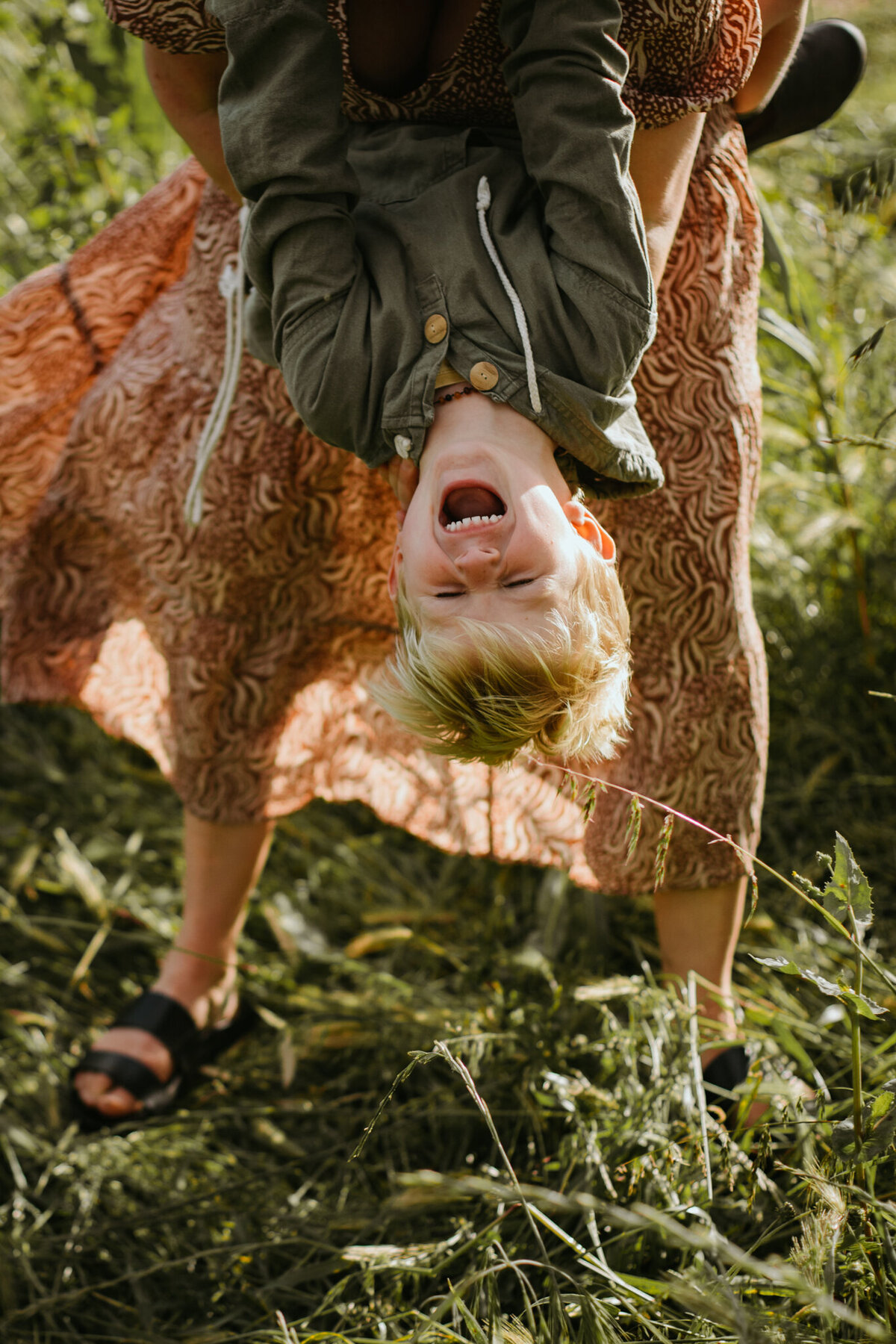 Mom’s legs visible as her son hangs upside down from her hips, laughing exuberantly.