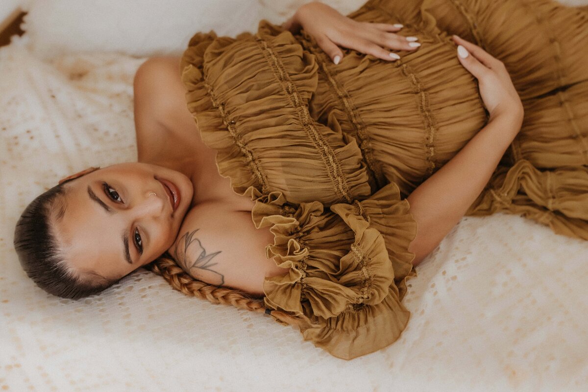 Person lying on a bed wearing a ruffled, off-shoulder brown dress. They have a tattoo on their upper arm and are looking directly at the camera.
