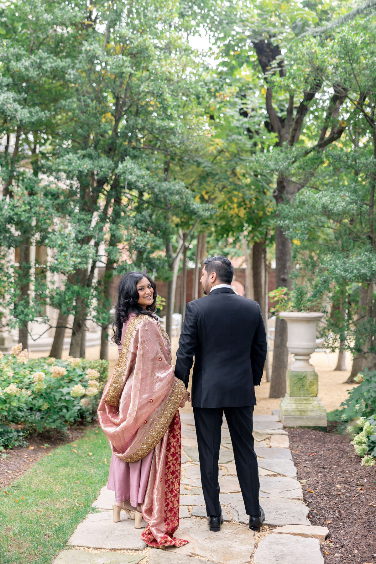 A couple stands on a stone path in a garden. The woman, wearing a pink and gold traditional outfit, looks over her shoulder. The man, dressed in a black suit, faces away. They are surrounded by lush green trees and shrubs.