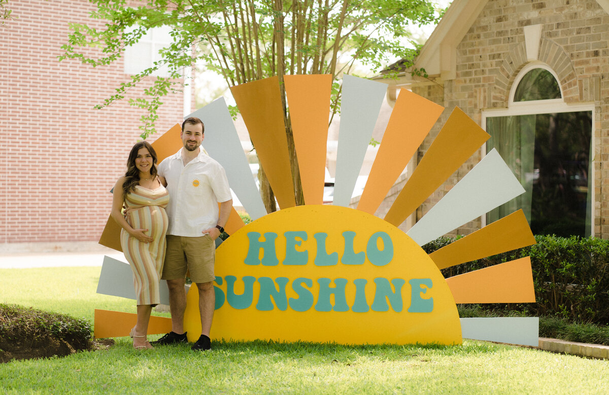 Man and pregnant woman posing in front of a Hello Sunshine baby shower backdrop design