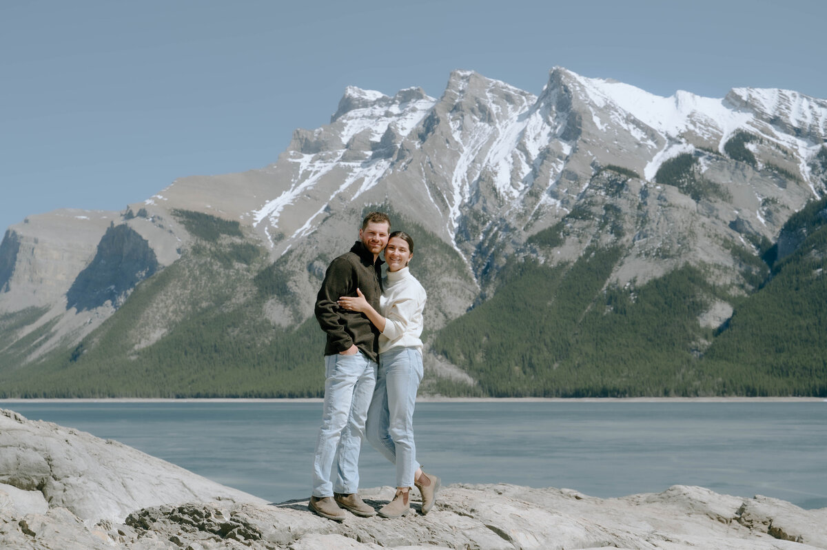 Engaged couple shares embrace with sweeping mountain view behind in Lake Minnewanka.