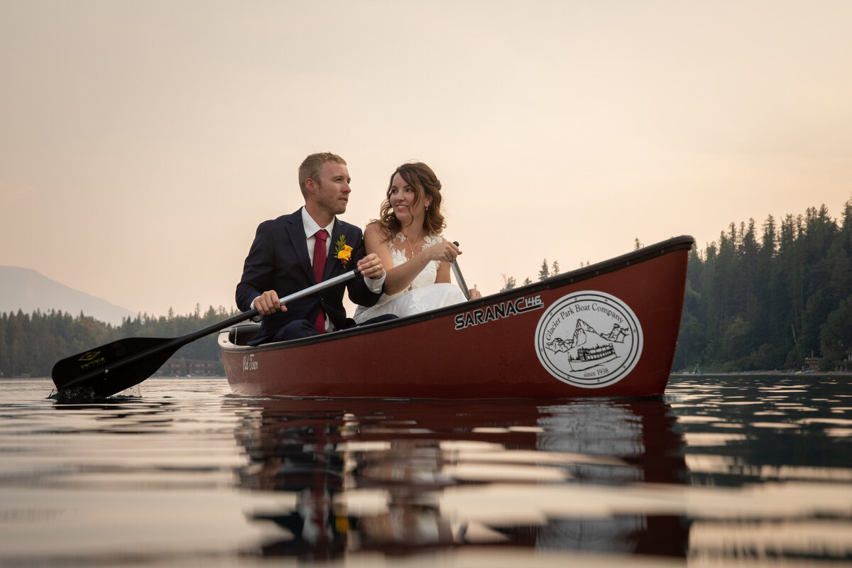 A bride and groom sit next to each other in a canoe on a lake in Glacier National Park, paddling through the water.