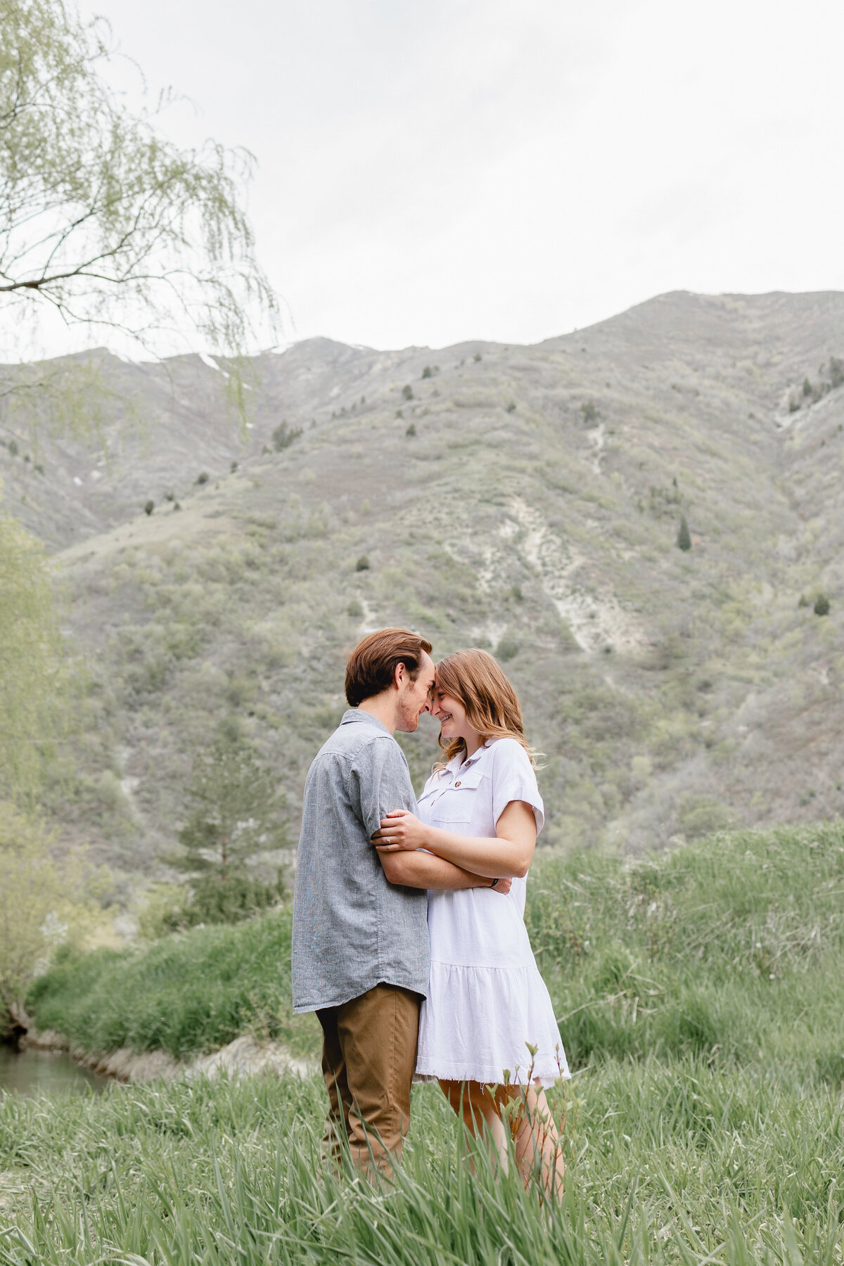 A couple stands close in a green meadow, touching foreheads with mountains behind them.