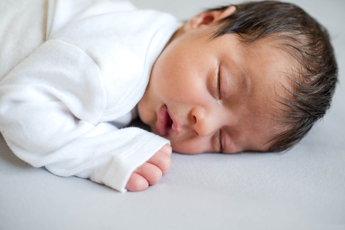 Newborn baby sleeping peacefully on their side, dressed in a white onesie.
