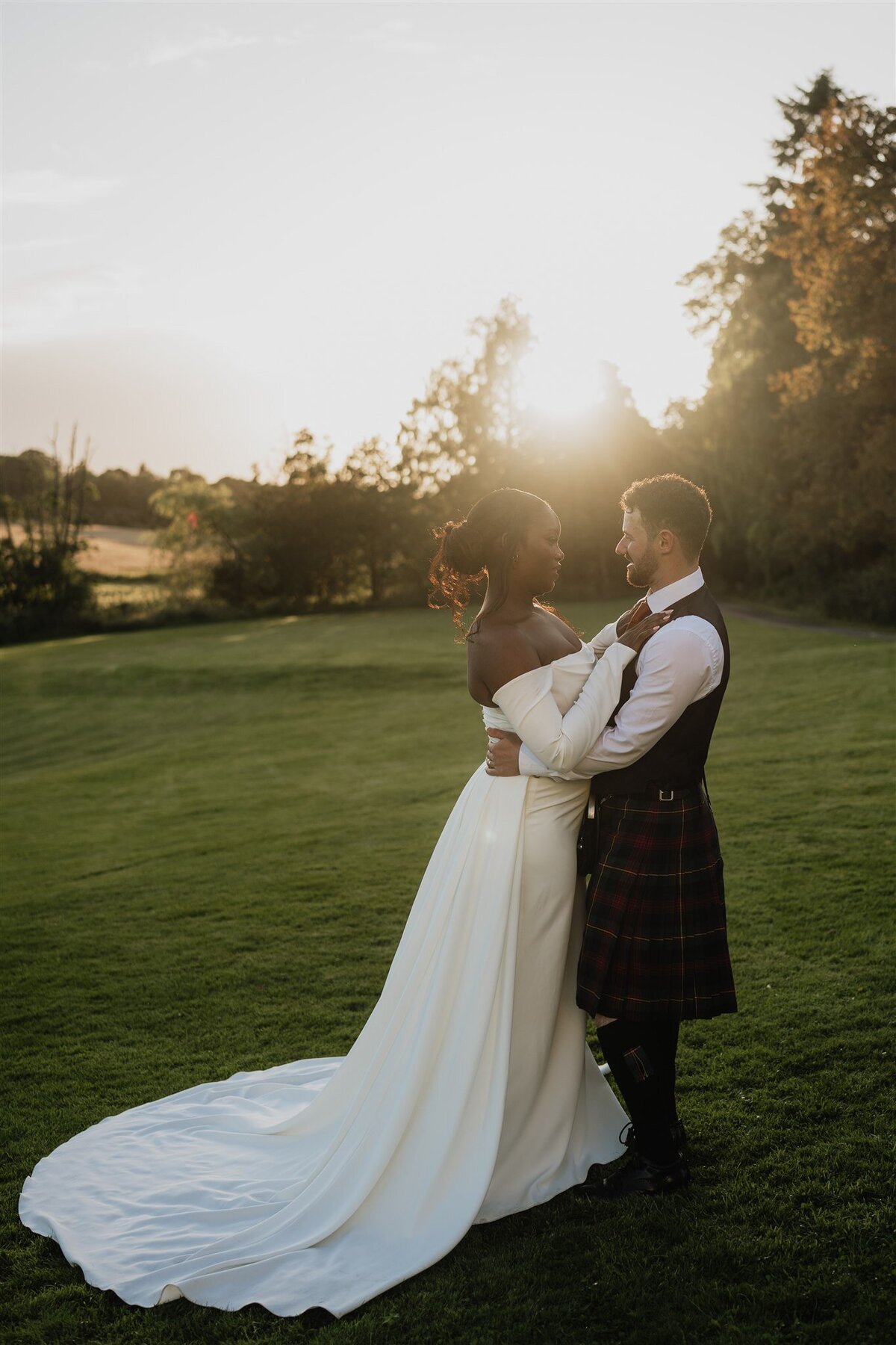 A wide photograph of a bride resting her hands of her husbands chest. Her wedding dress floats behind her and the sun sets between them. The stand in an open green space.
