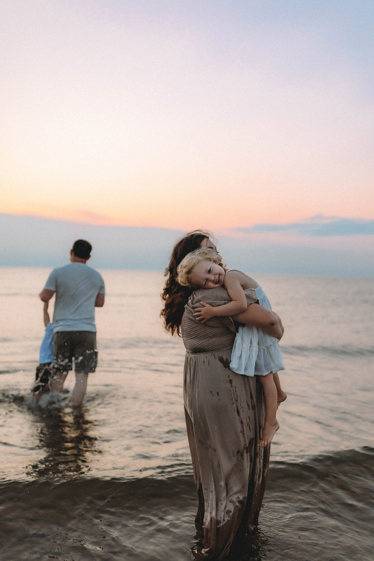 mom and daughter snuggle on the beach while dad and son play in background