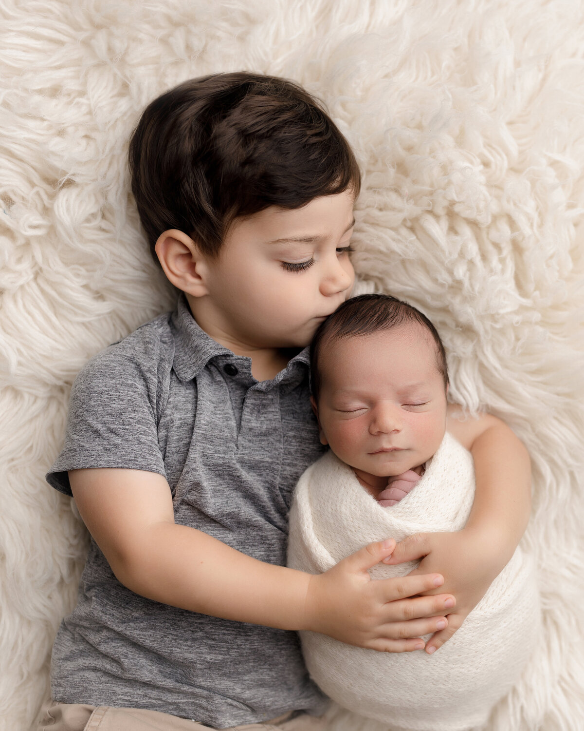 Newborn photoshoot in West Palm Beach and Jupiter photography studio. Big brother's arms are gently hugging baby brother atop of a fuzzy long faux fur rug. Baby's head is resting on big brother's shoulder and big brother is kissing the stop of his head. Baby is in a cream knit wrap with his fingers peeking out the top. Aerial image.