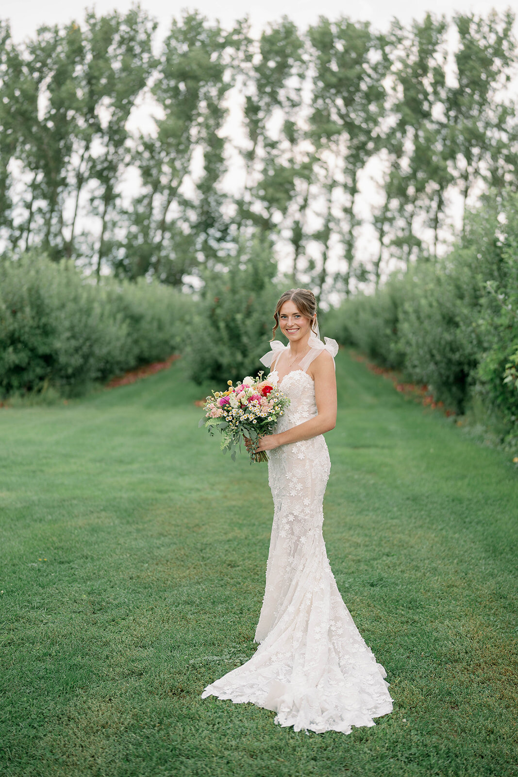 Full-body bridal portrait of the bride standing in front of the apple orchards during her September Cherry Barn wedding in Frankfort MI, featuring her lace gown and natural fall scenery.
