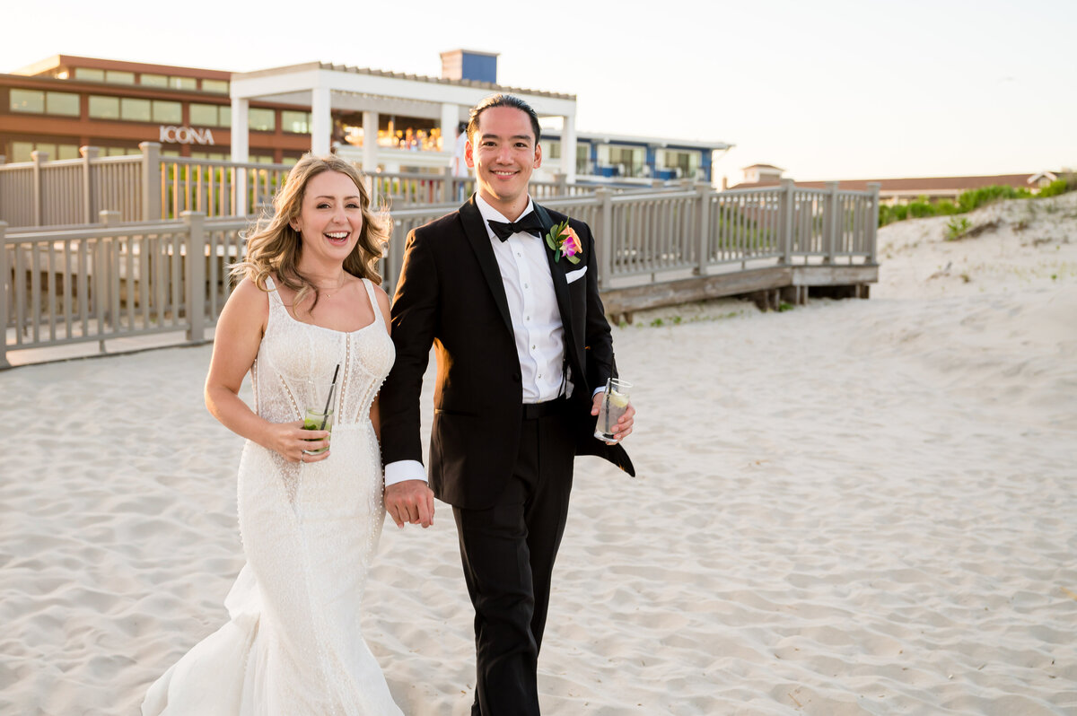 A bride and groom are walking on a sandy beach while holding hands, both smiling and holding drinks. The groom is in a black tuxedo, and the bride is wearing a white gown. Buildings are in the background.