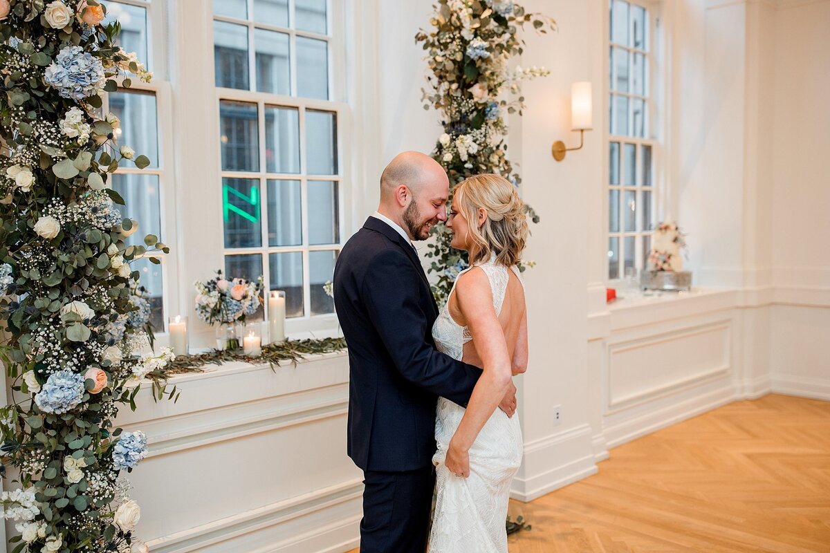 Bride wearing a backless halter top wedding dress embraces the groom wearing a dark navy suit at Noelle Nashville. The Saidee Ballroom is decorated with an  arbor of greenery accented with blue hydrangea, ivory roses and pink roses. The ballroom has tall windows and deep windowsills painted in ivory with blonde hardwood floors.