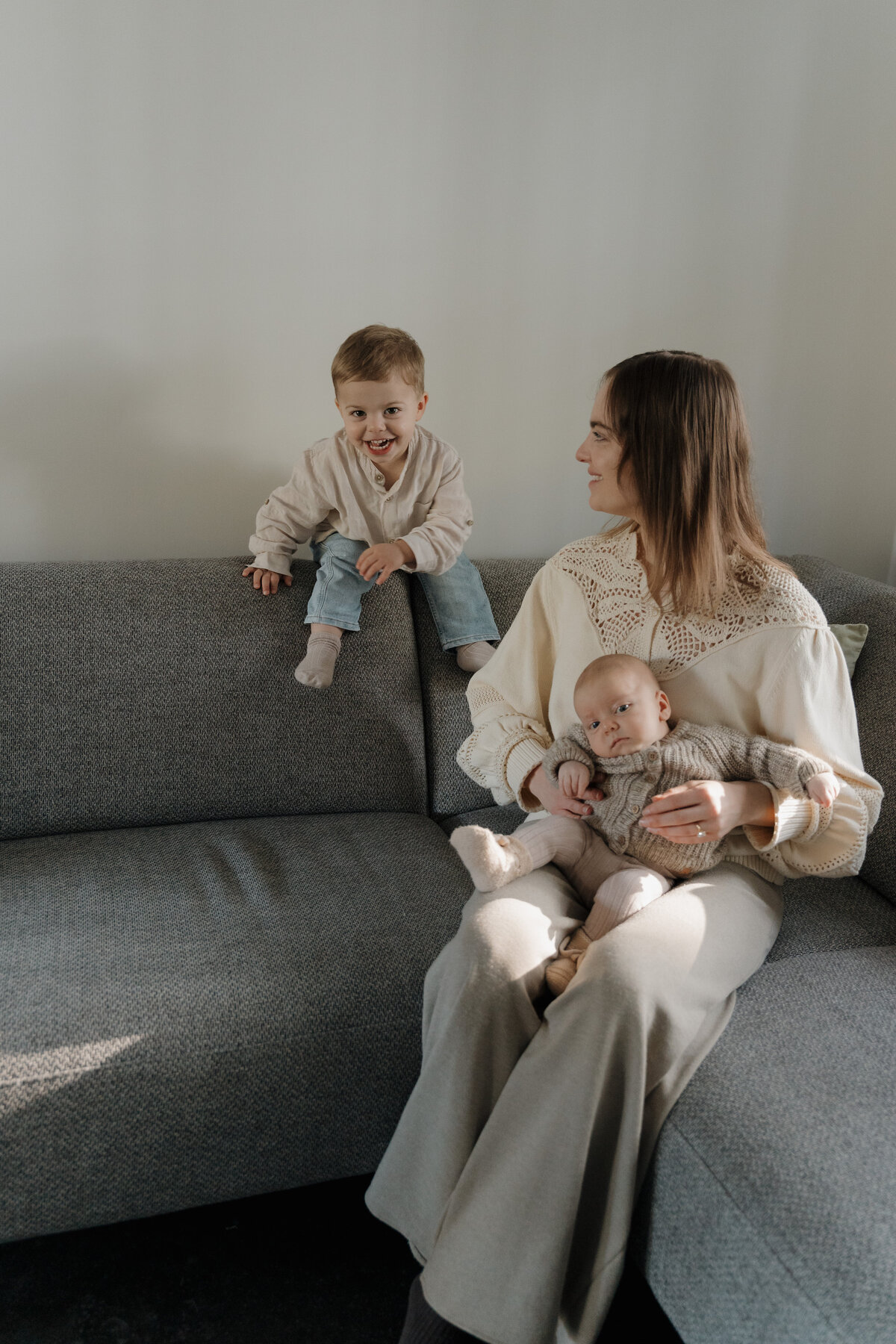 Moeder zit in de zetel met baby op haar schoot. Zoontje speelt in de zetel.