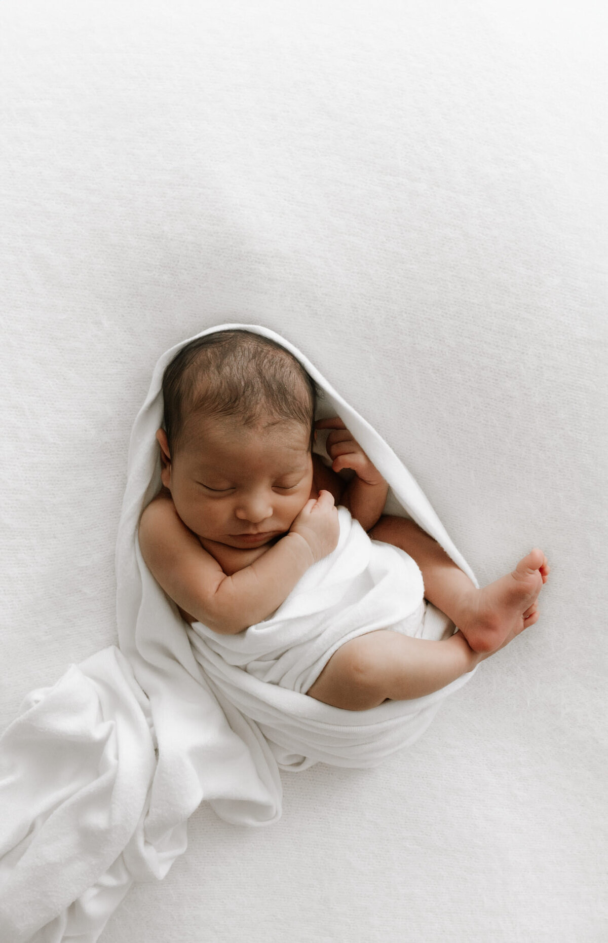 A serene newborn baby wrapped in soft white fabric, resting peacefully against a smooth white blanket. The minimalist setup and neutral tones highlight the baby's delicate features and curled-up position. Photographed in a Sydney newborn studio, this image captures the purity and calm of early days with a newborn.