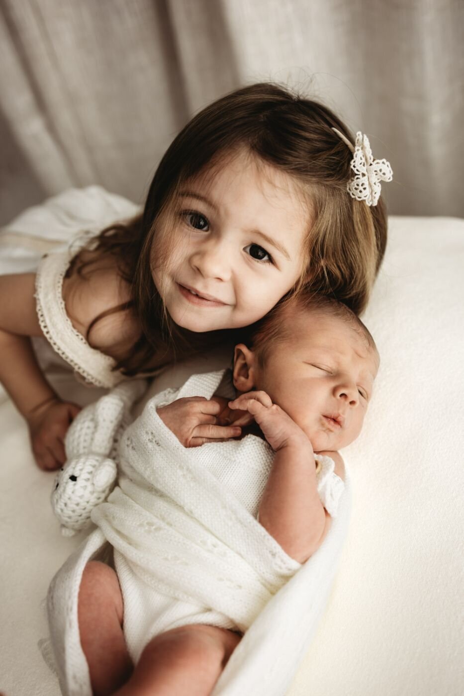 A smiling older sister leans over her newborn sibling, who is wrapped in a white knit blanket.