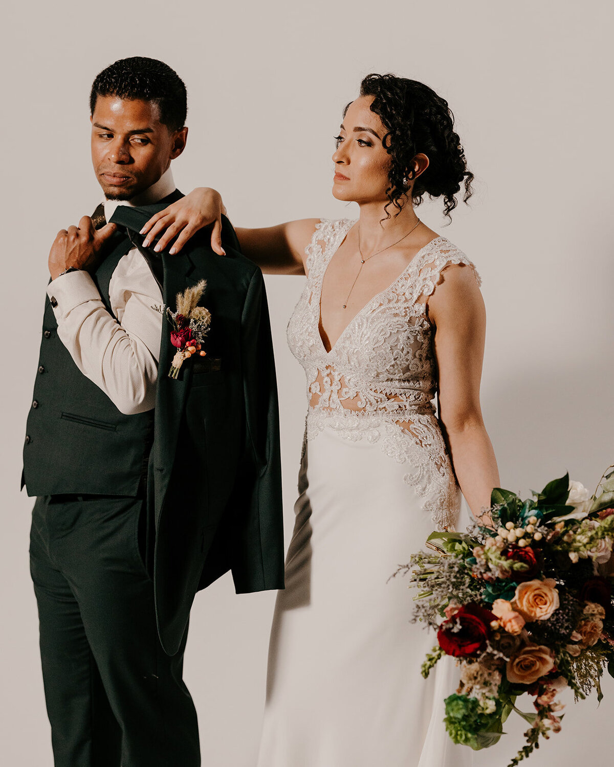 The bride holds a lush bouquet while the groom stands beside her with his jacket draped over his shoulder, both gazing off in opposite directions for a dramatic portrait.