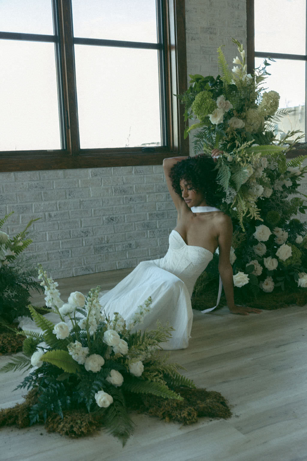 A person in a wedding dress sitting on the ground leaning against a floral arrangement.