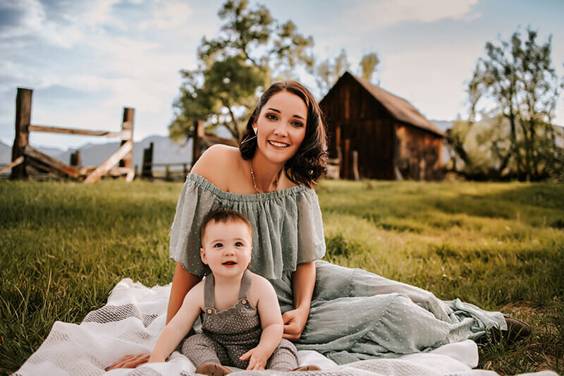 Mom wearing a sage green dress cuddles with infant son in knit overalls in Boulder Colorado in front of a brown barn