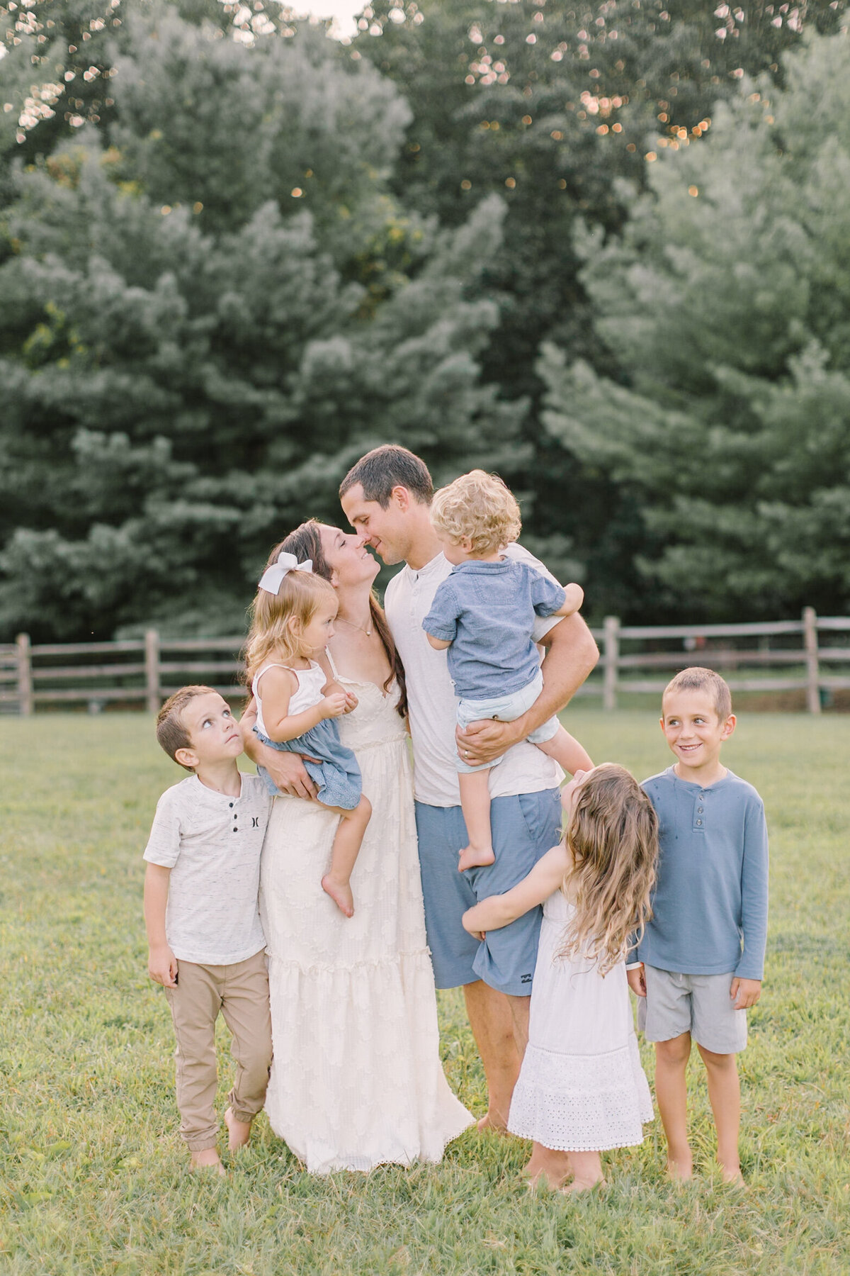 A family of six laughing together in a sunlit field, parents kissing while surrounded by their children — Portrait photography in Raleigh.