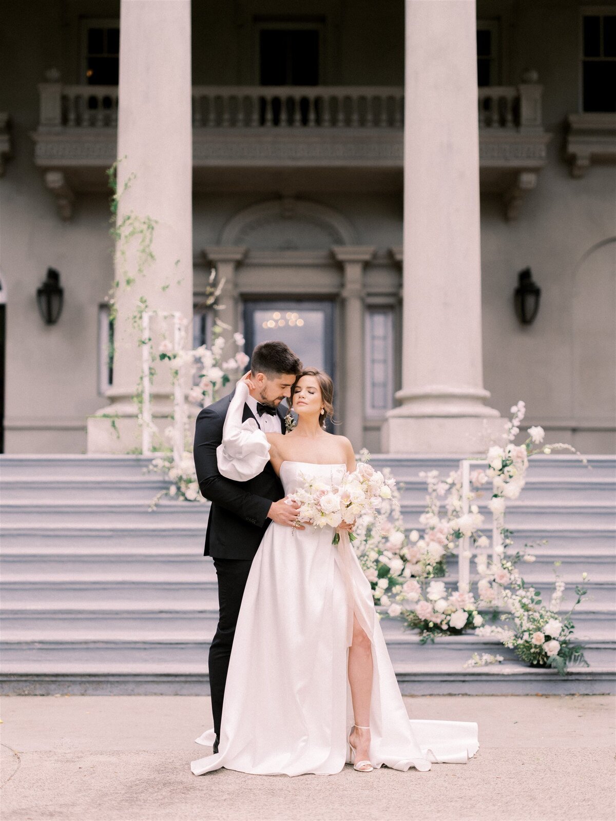 A bride and groom stand on steps in front of a building with columns. The groom embraces the bride from behind while she holds a bouquet of flowers. Their formal wedding attire is perfectly coordinated, thanks to their Canadian wedding planner's exquisite attention to detail.