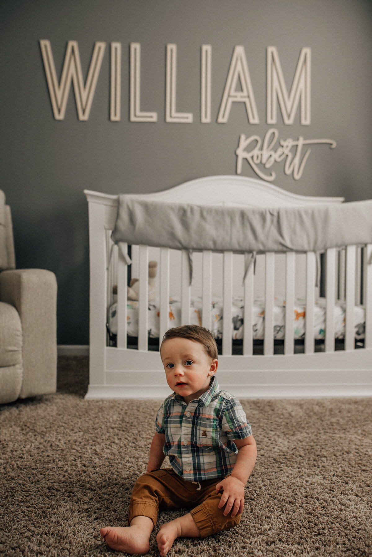 Young boy sits on the floor with a crib in the background.