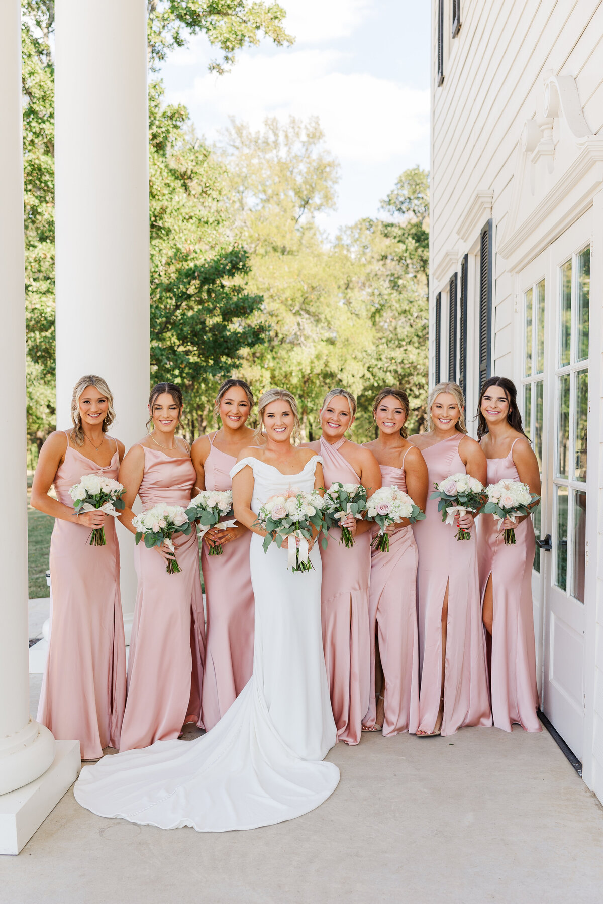 A joyful bride poses with her bridesmaids dressed in soft blush gowns at a stately wedding venue in Denton, Texas. Elegant outdoor bridal party portrait with a touch of Southern charm.
