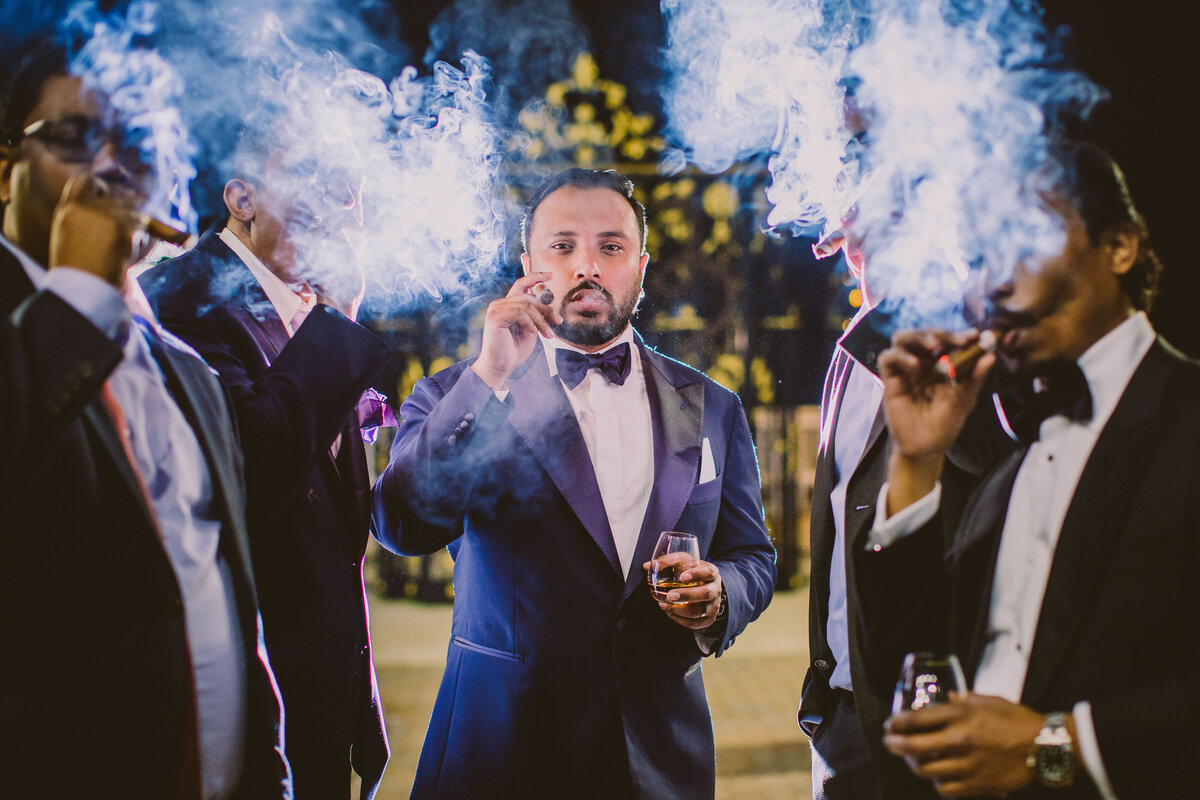 An indian groom smoking a cigar with his groomsmen outside at night at surf club ny