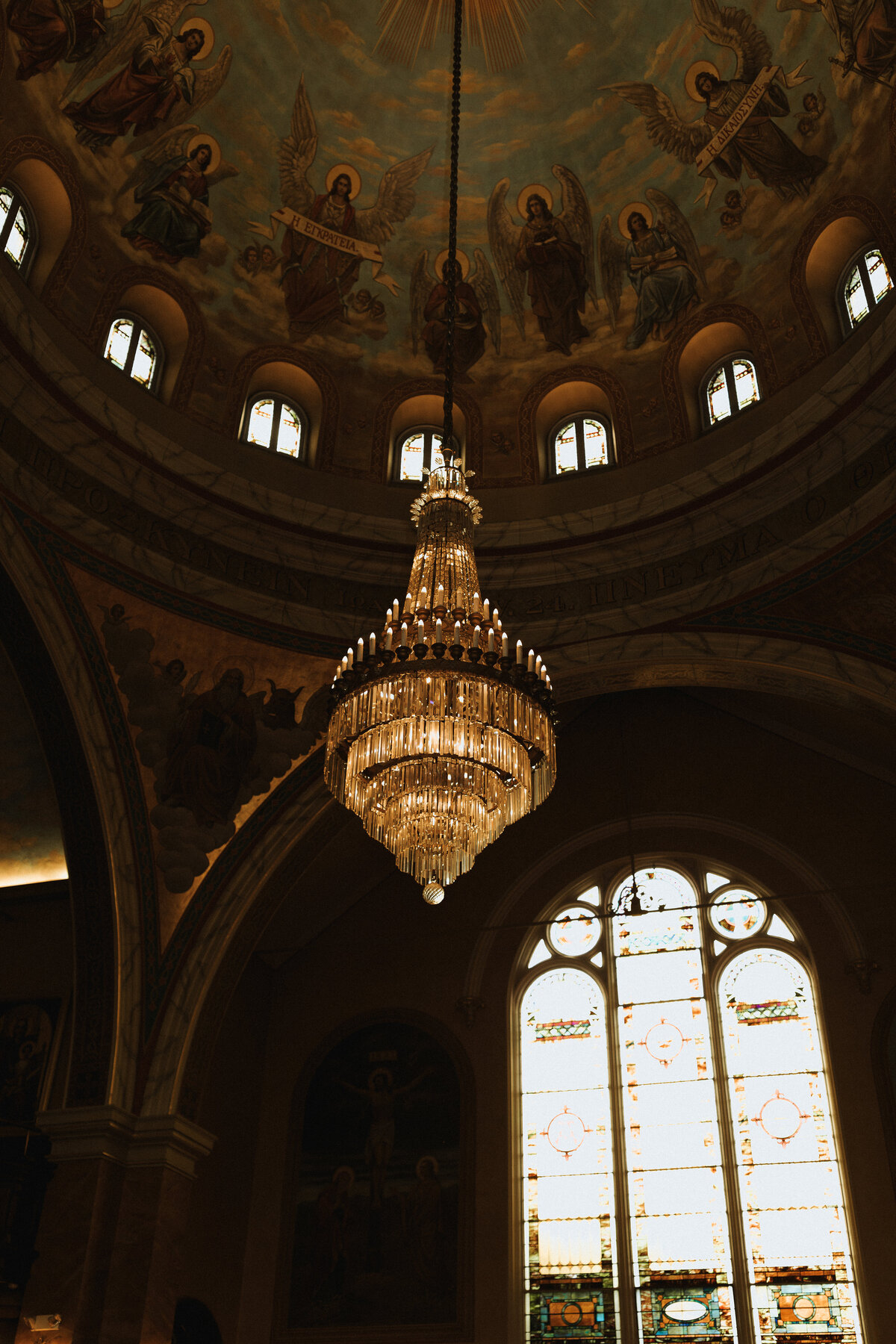 Ornate church interior featuring a crystal chandelier and painted dome ceiling with religious artwork and stained glass windows.