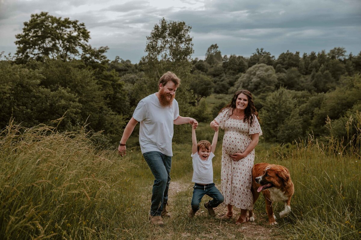 Expectant Mom, Dad, Toddler and Dog walking through Exeter, Ontario field. Toddler is swinging between mom and dad's arms, the dog is looking at the smiling toddler. Mom is in a long, causal dress holding her bump and smiling at the camera.