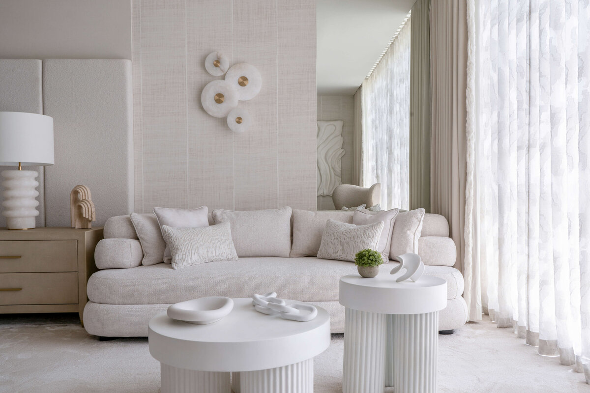 A white couch in the sitting area of the primary bedroom. Behind the couch is a large mirror and a circular white art piece.