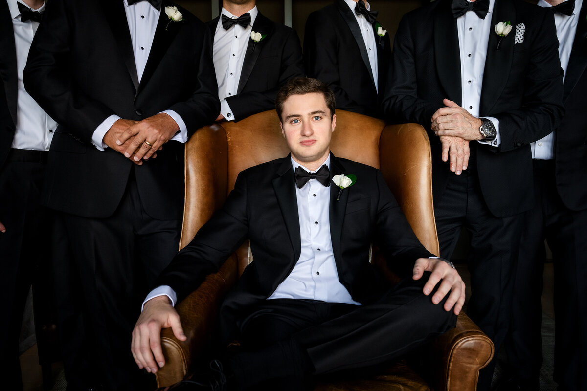 Husband to be sitting on a leather chair  surrounded by groomsmen